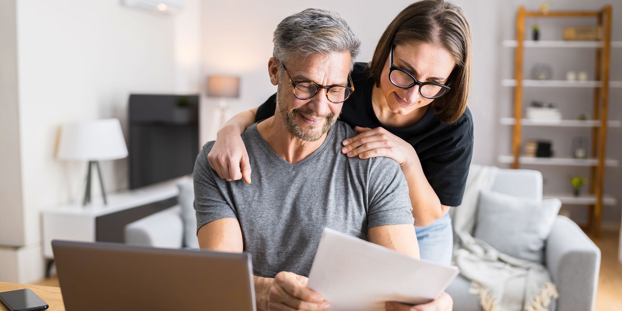 A woman leans over a man's shoulder as they smile at a financial document together.