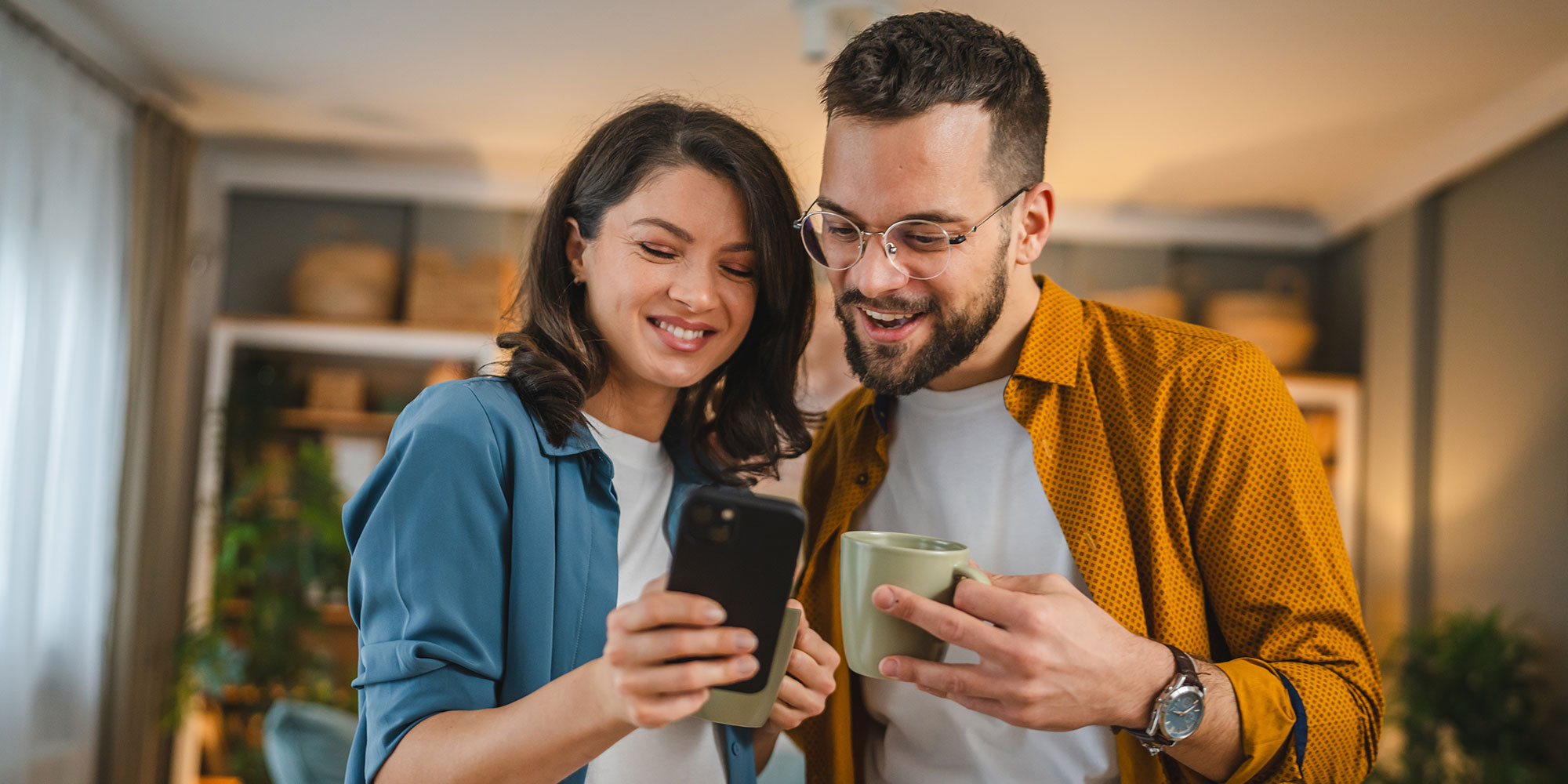 Man and woman each holding a mug and both looking at the woman's phone. 