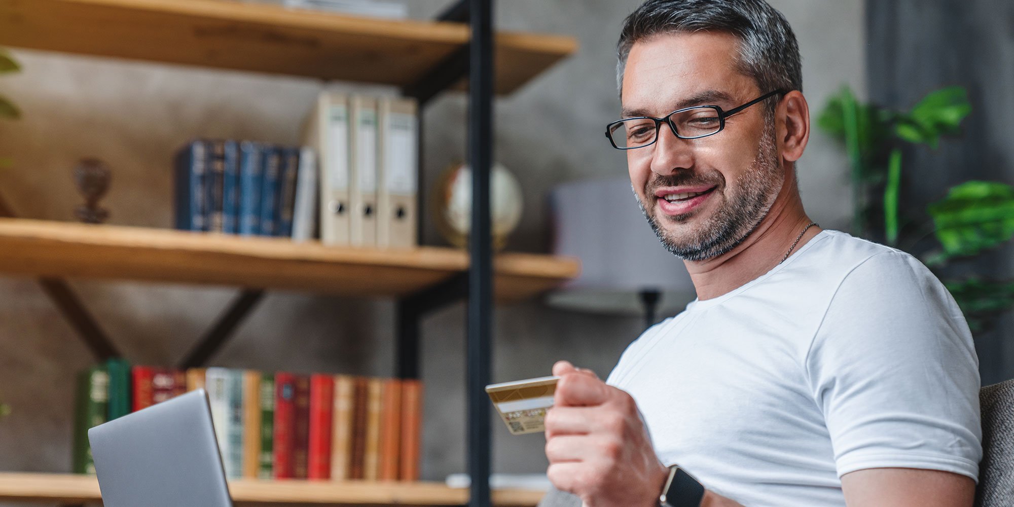 Middle aged man wearing glasses, holding up his credit card. 