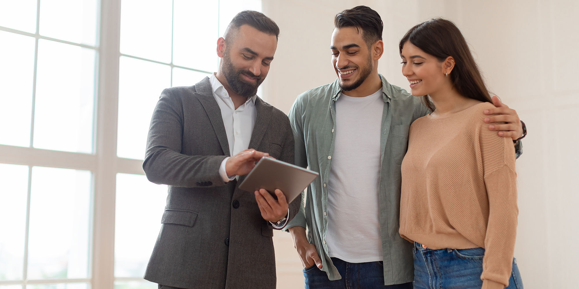 Real estate agent showing a property to a smiling couple using a tablet in a bright, spacious room.