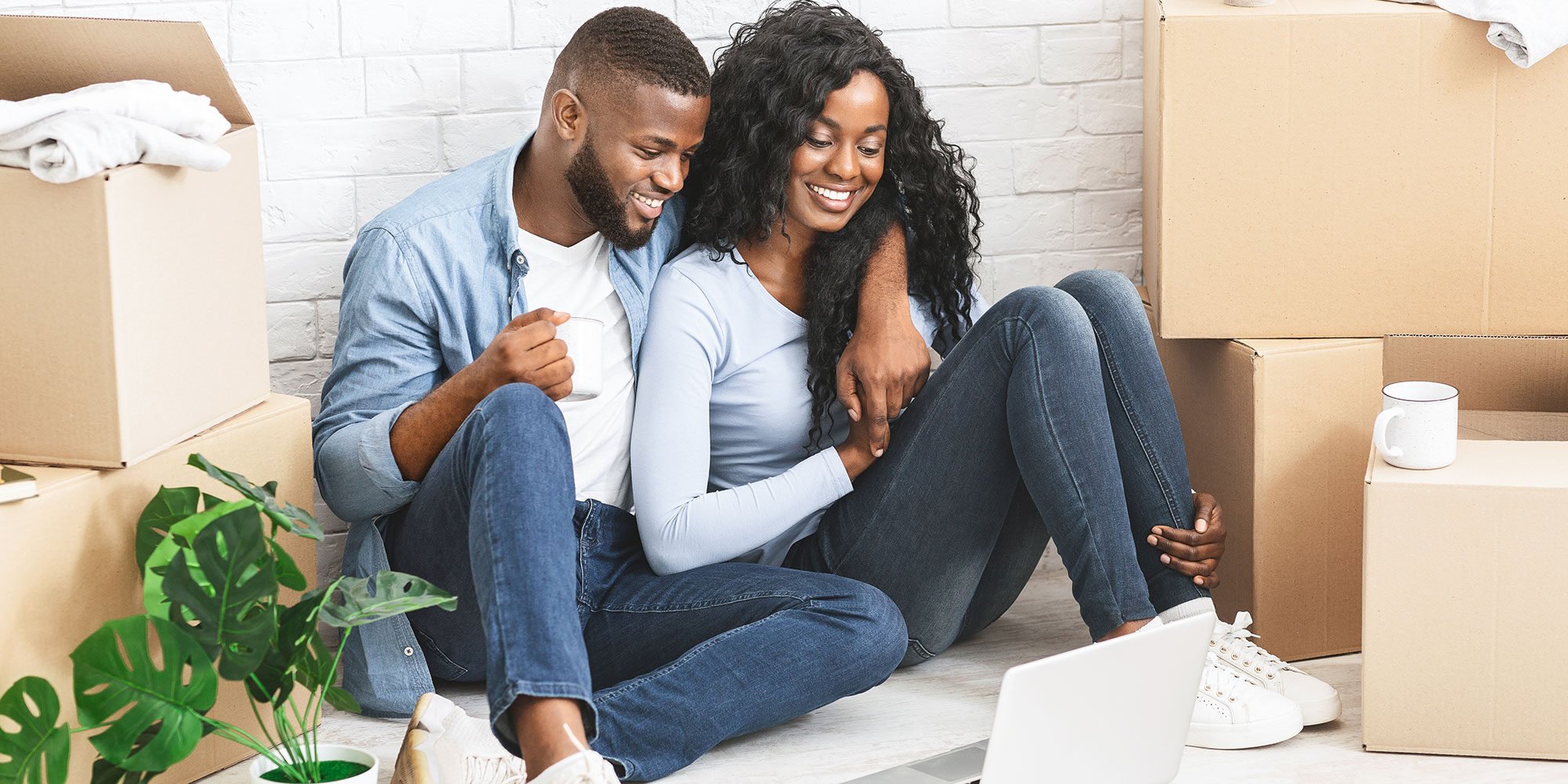 Happy couple sitting on the floor among moving boxes, looking at a laptop, in their new home