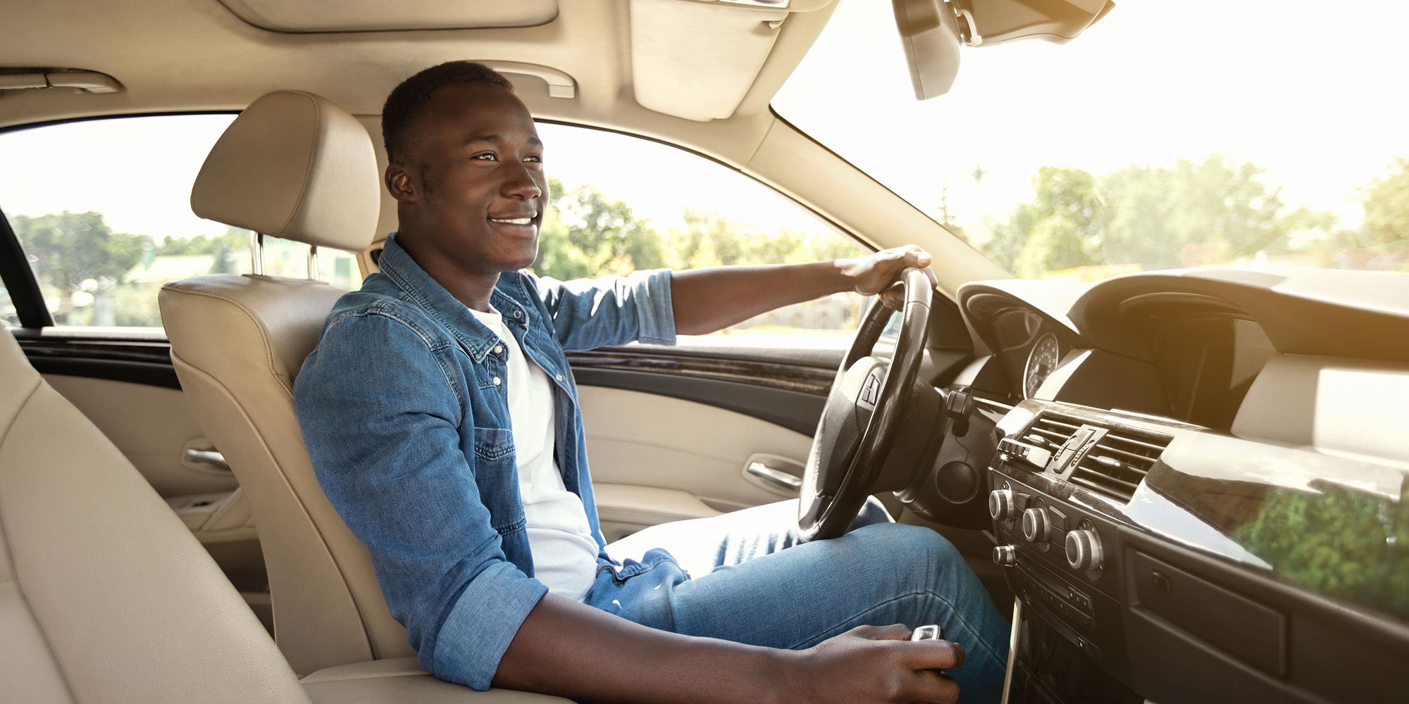 A young man smiling while driving a car on a sunny day. He is wearing a denim jacket and appears relaxed and confident, enjoying the drive with one hand on the steering wheel.